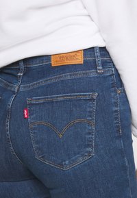 Jeans de denim azul oscuro con una etiqueta de cuero marrón en la cintura, que presentan un patrón de costura en forma de corazón en los bolsillos traseros y un detalle del logo en rojo.