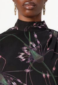 Blusa nera floreale con stampe botaniche verdi e rosa, colletto alto e dettagli a pieghe. Il materiale sembra leggero e strutturato.
