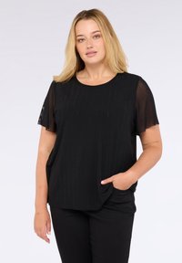 Donna bionda con pelle chiara che indossa una maglietta nera a maniche corte con maniche trasparenti e pantaloni neri, in piedi con una mano in tasca.