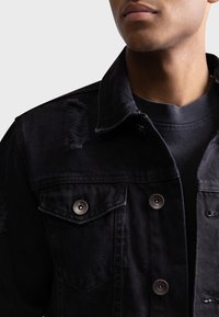 Chaqueta de mezclilla negra con acentos desgastados, dos bolsillos en el pecho con botones a presión metálicos y un cuello clásico. Llevada sobre una camisa oscura.