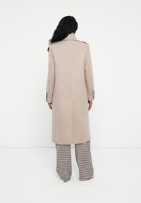 Cappotto beige in lana con design dritto, maniche lunghe e ventaglietto posteriore singolo. Accentuato con bottoni neri sui polsini. Indossato sopra pantaloni a quadri.
