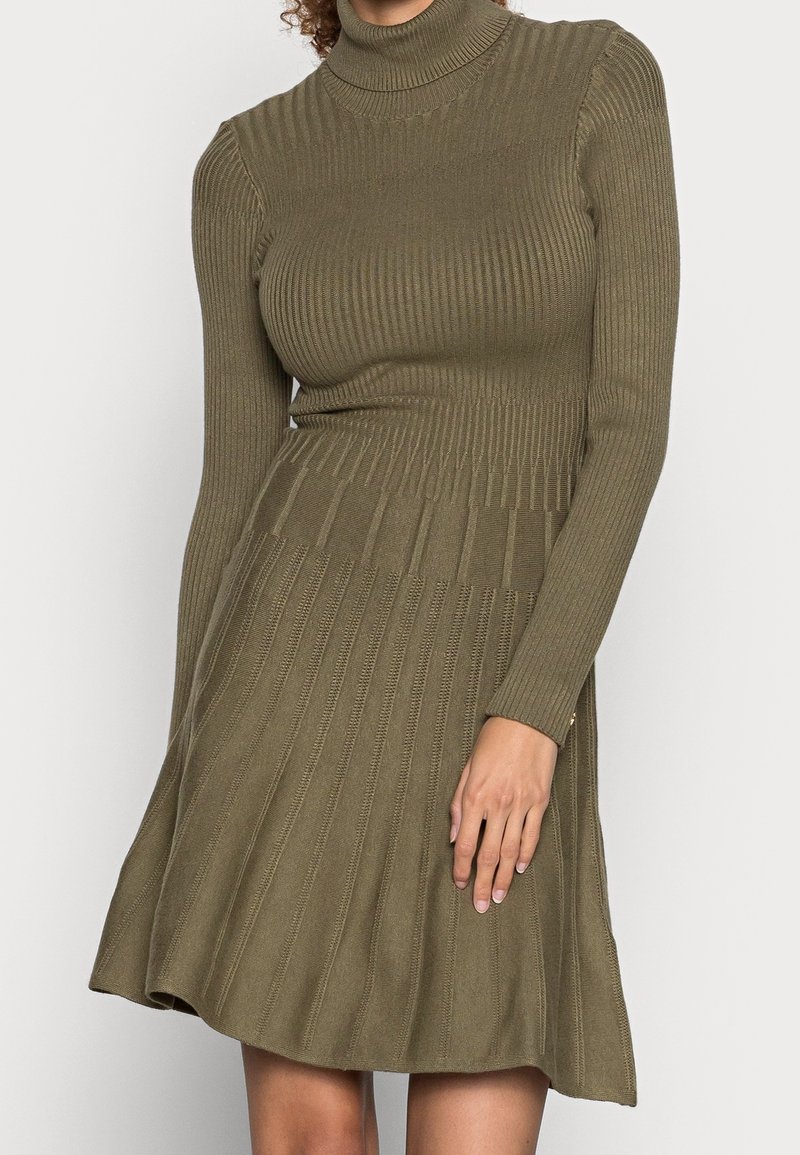 Femme portant une robe col roulé à manches longues côtelée en tricot vert olive avec jupe évasée, debout devant un fond neutre.