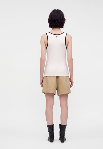 Personne aux cheveux courts et foncés, vue de dos, portant un débardeur blanc avec un bord noir, un short beige, et des bottes noires jusqu'au milieu du mollet.