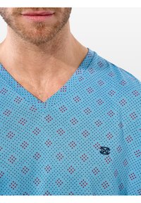 T-shirt azzurro chiaro a V con un motivo a pois composto da piccoli quadrati rossi. Il tessuto sembra morbido, con un logo ricamato sul petto.