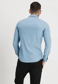 Camisa de mezclilla azul claro con mangas largas, puños con botones y dobladillo recto. Cuenta con cuello y un acabado de tela texturizada.