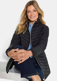 Smiling woman with long blonde hair sitting with legs crossed, wearing a black quilted jacket, blue sweater, and dark jeans.