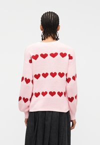 Άτομο με πλεγμένα μαλλιά  που φοράει ανοιχτοροζ πουλόβερ με κόκκινα μοτίβα καρδιάς στην πλάτη και στα μανίκια, συνδυασμένο με σκουρόχρωμη πλισέ φούστα.