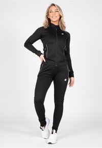 Chándal atlético negro hecho de poliéster con un diseño ajustado, chaqueta con cremallera y detalles blancos en los bolsillos y las mangas. Zapatillas blancas.