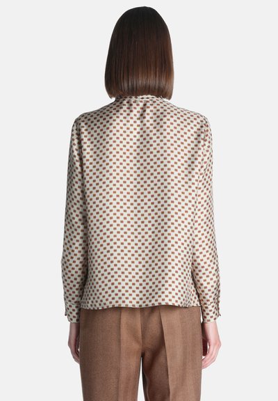 Femme aux cheveux bruns raides portant un chemisier crème à manches longues avec un motif géométrique orange et un pantalon marron taille haute, vue de dos.