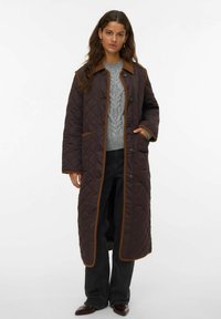 Lungo cappotto trapuntato marrone con un colletto marrone a contrasto, chiusura con bottoni, tasche laterali e un motivo a diamante sul tessuto.