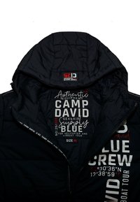 Chaqueta aislante negra con un patrón acolchado, que cuenta con capucha, etiquetas interiores con información, y detalles en contraste en blanco que indican la marca y la talla.