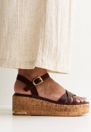 Pie con sandalia cuña de cuero marrón con tiras, plataforma de corcho y hebilla dorada, combinado con pantalones anchos de pierna texturizados en color blanco roto.