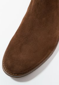 Bout de chaussure en daim marron avec une couture détaillée le long du bord et une semelle texturée visible sur un fond blanc.