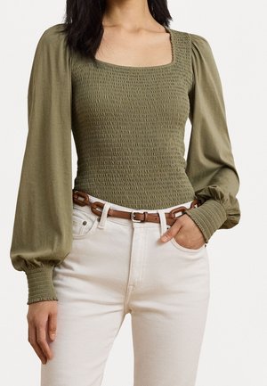 Nainen pukeutunut oliivinvihreään smokkattuun, neliökauluksiseen puseroon, jossa on pitkät liehukkeet hihanpäissä, ja valkoisiin korkeavyötäröisiin housuihin, joissa on ruskea vyö.