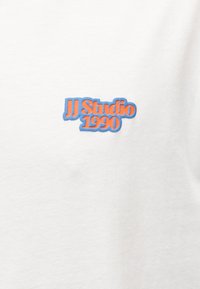 Λευκό ύφασμα με ένα μικρό λογότυπο που αναγράφει «JJ Studio 1990» με πορτοκαλί γράμματα περιγραμμένα σε μπλε, στο κέντρο της περιοχής του στήθους.