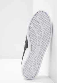 Semelle de basket noire et grise avec une semelle extérieure en caoutchouc blanc texturé, présentant un motif de galets et un logo sur la semelle intermédiaire.