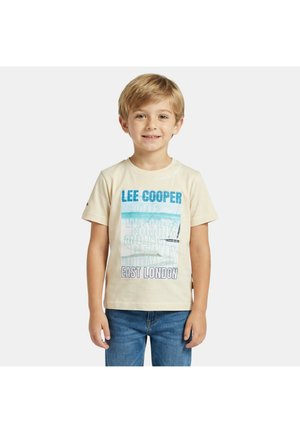 Jeune garçon souriant, portant un t-shirt beige graphique avec les textes "LEE COOPER" et "EAST LONDON", associé à un jean bleu, debout devant un fond uni.