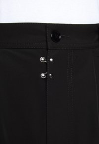 Pantalones de tela negra con una textura suave, que cuentan con un cierre de botón y dos broches metálicos con detalles de costura en el frente.