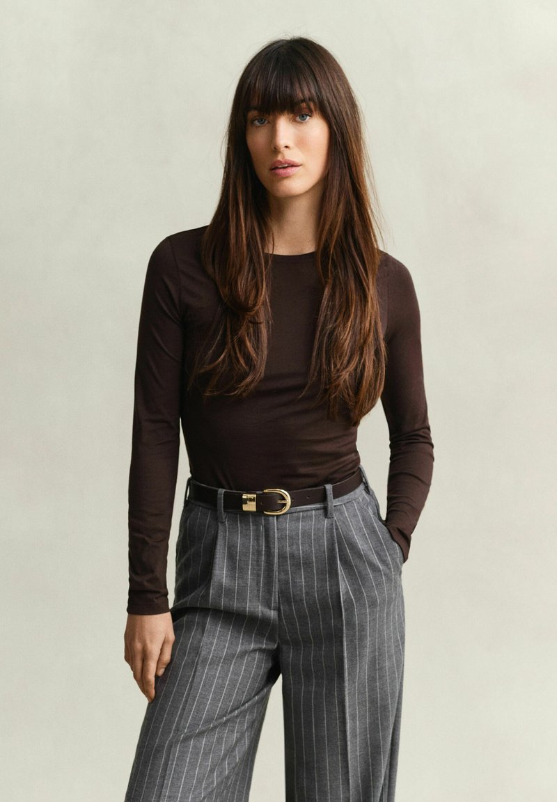 Femme aux longs cheveux bruns portant un haut marron foncé à manches longues et un pantalon gris à fines rayures avec une ceinture noire, debout devant un fond clair uni.