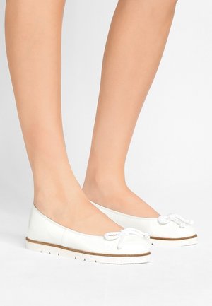 Pieds portant des ballerines blanches avec un détail de nœud en corde et des semelles blanches texturées, debout sur un fond clair uni.