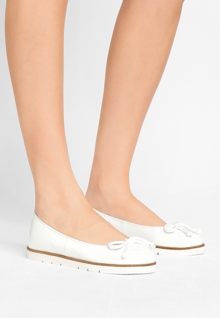 Pieds portant des ballerines blanches avec un détail de nœud en corde et des semelles blanches texturées, debout sur un fond clair uni.