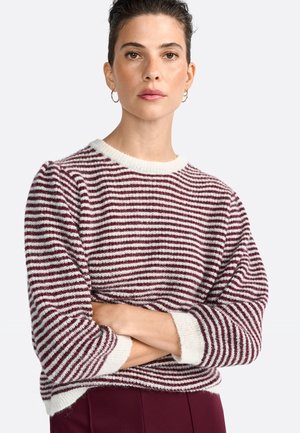 Una donna con capelli corti e scuri indossa un maglione a righe bordeaux e bianche, con le braccia incrociate, contro uno sfondo semplice. Ha un'espressione sicura di sé.