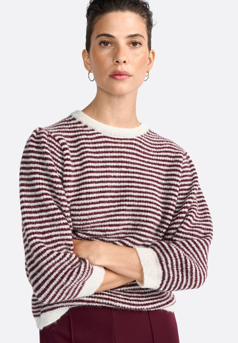 Una donna con capelli corti e scuri indossa un maglione a righe bordeaux e bianche, con le braccia incrociate, contro uno sfondo semplice. Ha un'espressione sicura di sé.