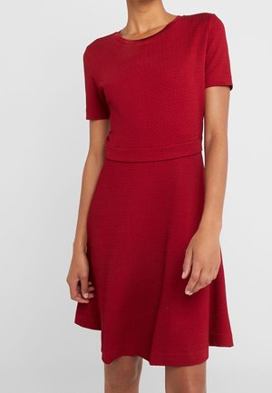 Femme portant une robe rouge texturée, courte aux manches, longueur au genou, taille cintrée et encolure ronde sur un fond clair uni.