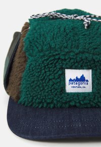 Kapica za vanjske aktivnosti s panelima od zelene i smeđe flis tkanine, donjim dijelom u mornarsko plavoj boji, detaljem od užeta i logotipom Patagonia na prednjoj strani.