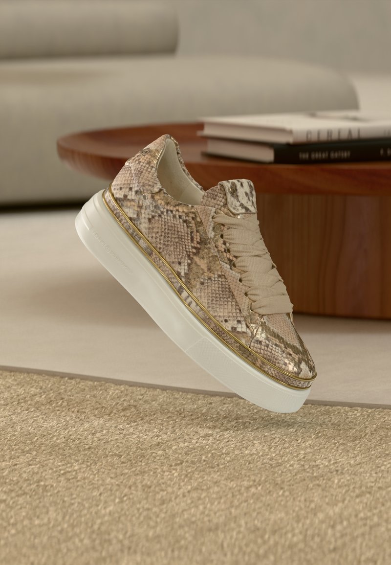 Αθλητικό παπούτσι με σχέδιο φολιδωτής επιδερμίδας σε μπεζ και καφέ χρώμα, με παχιά λευκή σόλα και μπεζ κορδόνια, δίπλα σε στρογγυλό ξύλινο τραπέζι και βιβλία πάνω σε μπεζ χαλί.