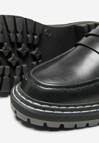 Zapatos de cuero negro con punta redondeada, que presentan una suela de goma gruesa con un patrón de agarre texturizado y costuras blancas a lo largo del borde.