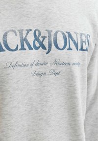 Sweat-shirt gris avec texte bleu marine "JACK & JONES" et texte plus petit en dessous. Le tissu semble doux, avec une texture lisse.