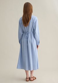 Vestito azzurro chiaro a maniche lunghe con righe verticali e vita con fiocco. Il tessuto sembra morbido e l'orlo arriva a metà polpaccio.