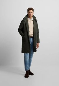Abrigo exterior verde con capucha, llevado sobre un suéter beige, combinado con vaqueros de un azul claro y botas de tobillo marrón oscuro. Diseño sencillo, con botones en la parte delantera.