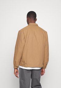 Chaqueta de algodón marrón con cuello, detalles de costura visibles, bolsillos laterales y cierres de presión en los puños, vista desde la parte trasera.