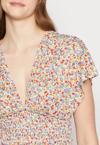 Robe à motif floral avec un décolleté en V, des manches courtes et une taille smockée. Présente de petites fleurs rouges, bleues et jaunes sur un fond clair.