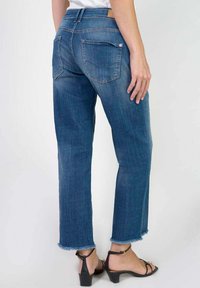 Jean droit en denim bleu avec un ourlet effiloché, présentant des poches arrière et une taille haute. Porté avec des sandales noires à brides.