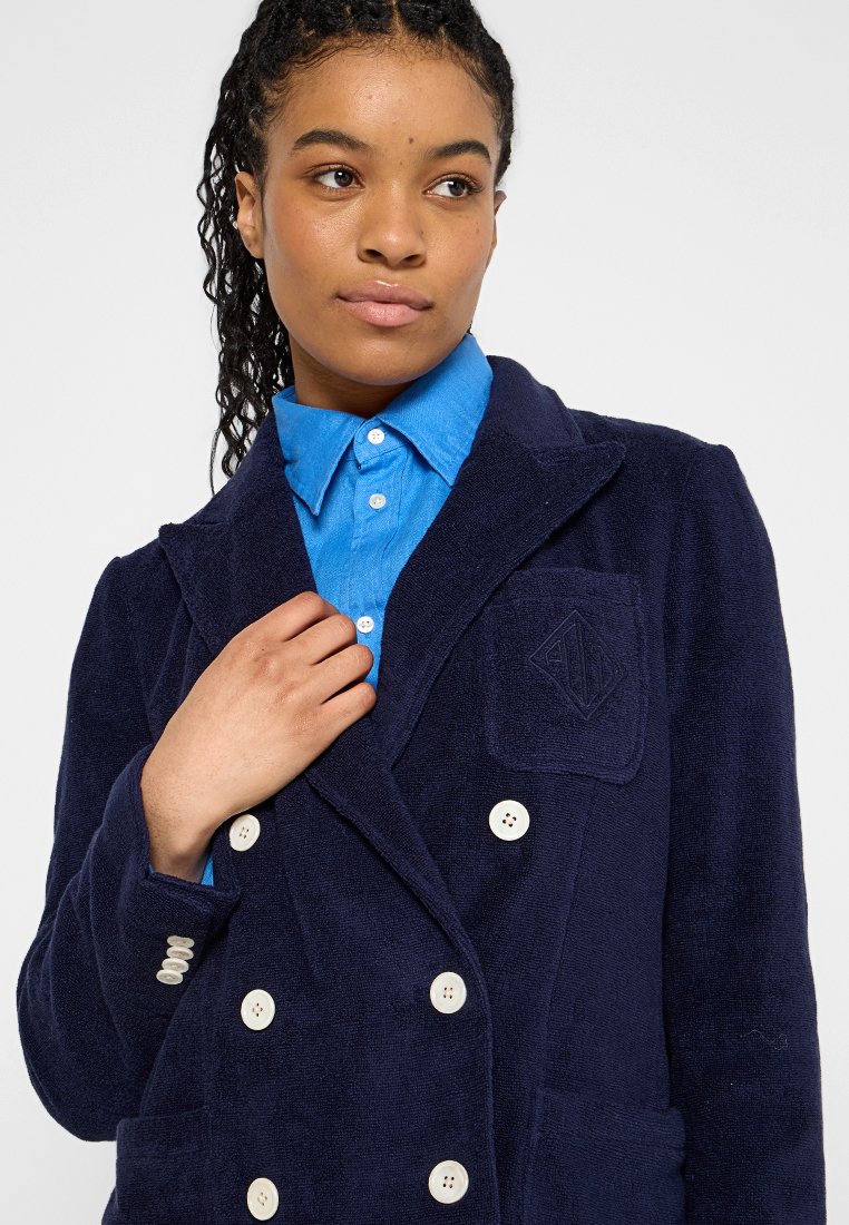 Kobieta z warkoczami, ubrana w niebieską koszulę z guzikami pod granatowym podwójnym płaszczem, trzymająca po jednym ręku kołnierz płaszcza.