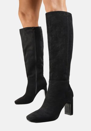 Bottes noires jusqu'au genou en suède doux, avec un bout pointu et un talon épais au fini brillant. Fermeture éclair sur le côté pour un enfilage facile.