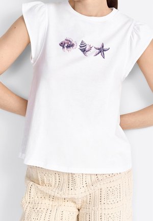 Biały krótki top z krótkim rękawem z fioletowym wzorem muszli morskiej i rozgwiazd, noszony z kremowymi, dzianinowymi szortami.