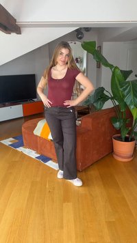 Νεαρή γυναίκα με μακριά μαλλιά στέκεται μέσα σε εσωτερικό χώρο κοντά σε πορτοκαλί καναπέ και μεγάλο πράσινο φυτό, φορώντας βυσσινί μπλούζα, σκούρο παντελόνι και λευκά παπούτσια.