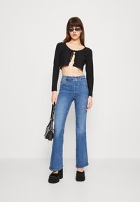 Top nero a maniche lunghe e a croppato con nodo frontale, abbinato a jeans flare blu a vita alta. La modella indossa scarpe con platform e una piccola borsa nera.
