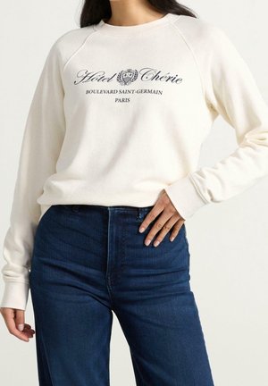 Femme portant un pull crème avec le texte "Hotel Chérie Boulevard Saint-Germain Paris" et un jean taille haute bleu foncé, main sur la hanche.