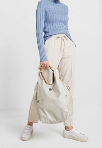 Person iført en lyseblå ribbet sweater, beige drawstring bukser, hvide sneakers, som holder en stor offwhite tote bag med brune håndtag.