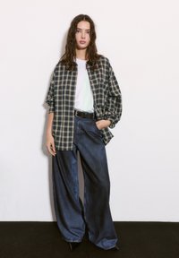 Cămașă supradimensionată cu carouri în nuanțe de bleumarin și crem, asociată cu blugi întunecați, cu croială lejeră. Modelul poartă un tricou alb și o curea neagră. Aspect casual, elegant.