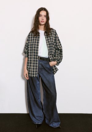 Koszula w kratę w kolorze granatowym i kremowym w oversize'owym fasonie, zestawiona z szerokimi dżinsami w ciemnym kolorze. Modelka ma na sobie białą koszulkę i czarny pasek. Gładki, casualowy wygląd.