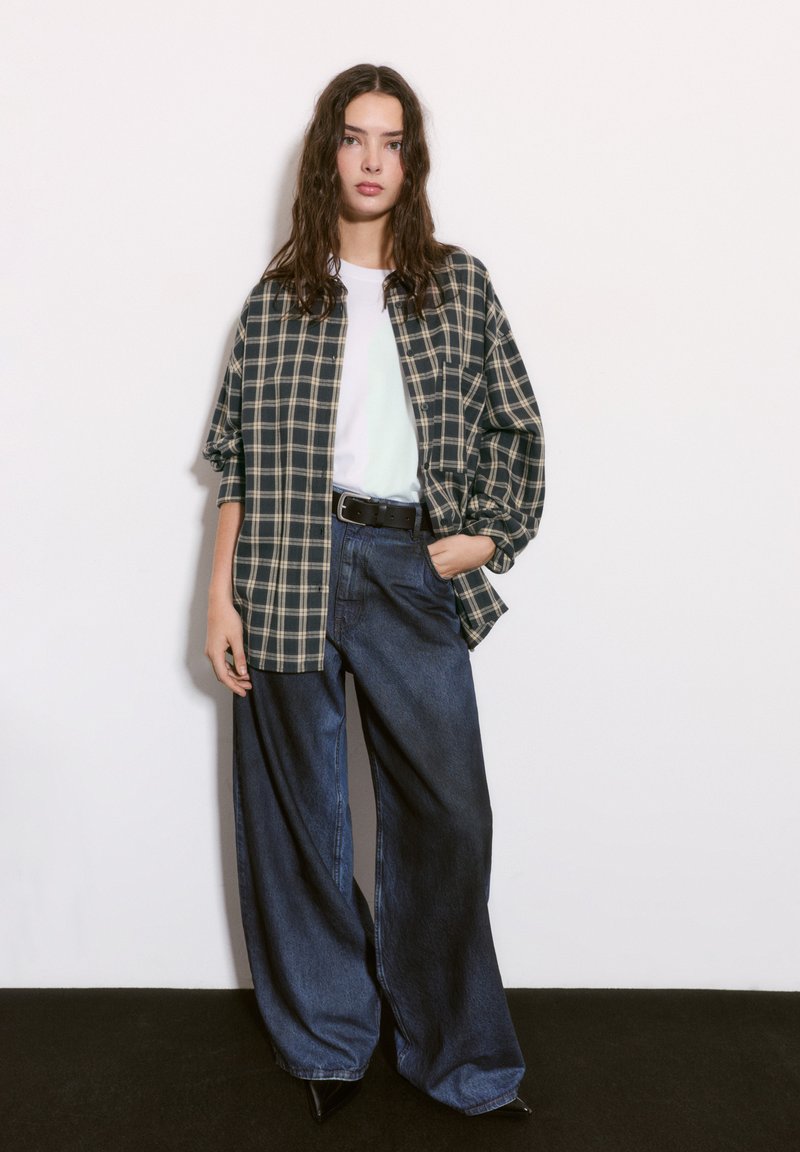 Cămașă supradimensionată cu carouri în nuanțe de bleumarin și crem, asociată cu blugi întunecați, cu croială lejeră. Modelul poartă un tricou alb și o curea neagră. Aspect casual, elegant.