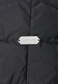 Black puffer jacket fabric with diagonal stitching, featuring a silver metal tag engraved with "QUARTZ CO." Smooth texture, lightweight design.