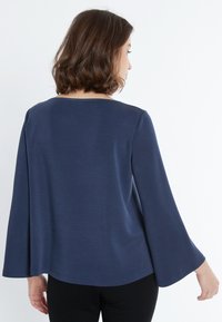 Dunkelblaues Oberteil mit Glockenärmeln, glatter Textur, runden Ausschnitt und lockerer Passform, kombiniert mit schwarzen Hosen. Rückansicht gezeigt.