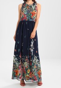 Kvinna i ärmlös mörkblå maxiklänning med färgglad blommönster på överdelen och fållen, i kombination med röda klackar med öppen tå.